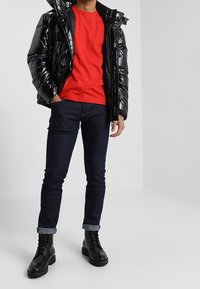 Osoba v lesklé černé péřové bundě, červeném tričku, tmavých ohrnutých džínách a černých šněrovacích botách stojící na jednobarevném pozadí.