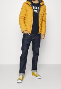 Gul puffjacka med huva, dragkedjestängning, mörkblå t-shirt under, mörka jeans och gula sneakers med vita sula och snören.