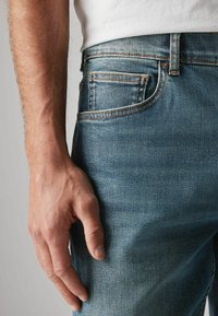 Jeans de mezclilla azul oscuro con un diseño ajustado, que cuentan con un estilo de cinco bolsillos y costuras en contraste. Mano descansando en el costado.
