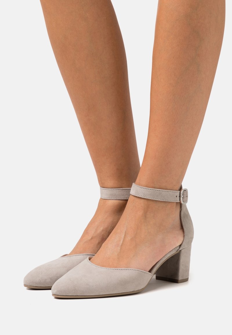 Talons carrés en daim beige avec brides à la cheville fixées par de petites boucles sur les pieds, sur fond uni.