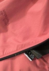 Rosa Jackenstoff mit teilweise geöffnetem schwarzem Reißverschluss, der das Jack Wolfskin-Logo und ein schwarzes Pflegeetikett im Inneren zeigt.