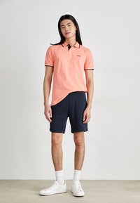 Rožinė polo marškinė su juodu apvadu, tamsiai mėlynos šortai ir balti kedai. Modelis stovi neutraliame fone, demonstruodamas kasdienį aprangą.