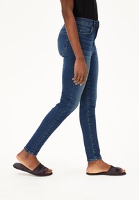 Skinny-Jeans aus dunklem Denim mit mittelhoher Taille, dezenten Verwaschungen und klassischem Fünf-Taschen-Design, kombiniert mit schwarzen Riemchensandalen.