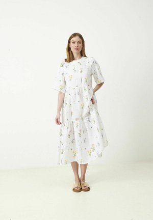 Nainen seisoo valkoista taustaa vasten valkoisessa midi-mekossa, jossa on värikkäitä kukkakuvioita, ja beiget sandaalit jalassa, toinen käsi taskussa.