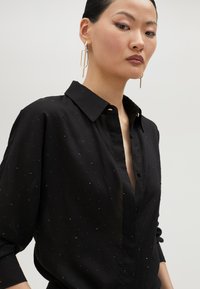 Black button-up shirt with a collared neck and long sleeves, featuring a textured pattern of small raised dots. Smooth fabric and fitted design.