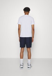 Mies seisoo selkä kameraan, päällään valkoinen Lacoste-T-paita, tummansiniset shortsit, valkoiset sukat ja valkoiset lenkkarit yksinkertaista valkoista taustaa vasten.