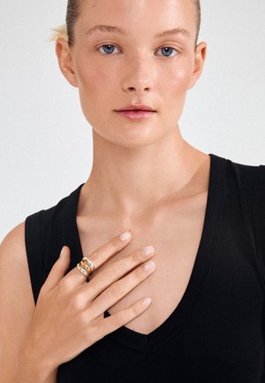 Femme aux yeux bleus portant un haut noir sans manches touche sa poitrine de la main gauche, montrant des bagues en or et en argent sur ses doigts.