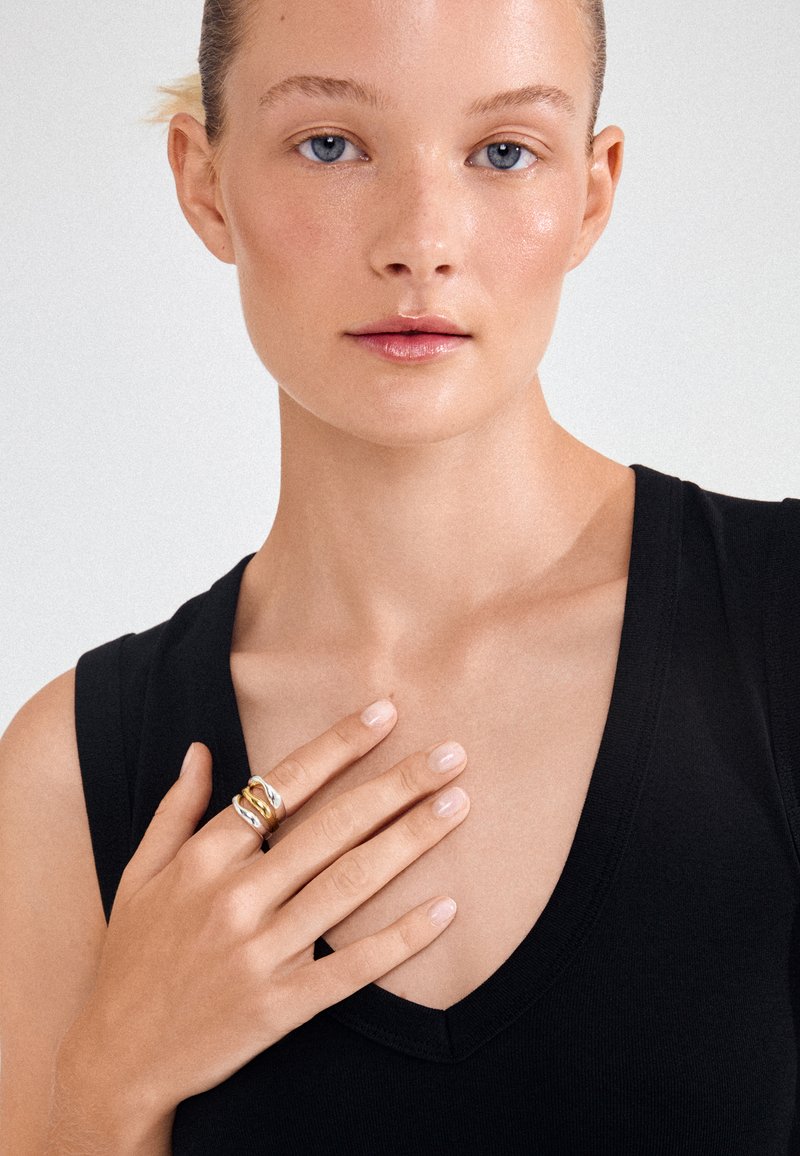 Mujer con ojos azules que lleva una camiseta negra sin mangas toca el pecho con la mano izquierda, mostrando anillos de oro y plata en los dedos.