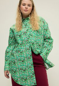 Blusa a maniche lunghe con colletto, verde a fantasia, e bottoni. Il design presenta forme astratte in tonalità di verde, rosso e nero.