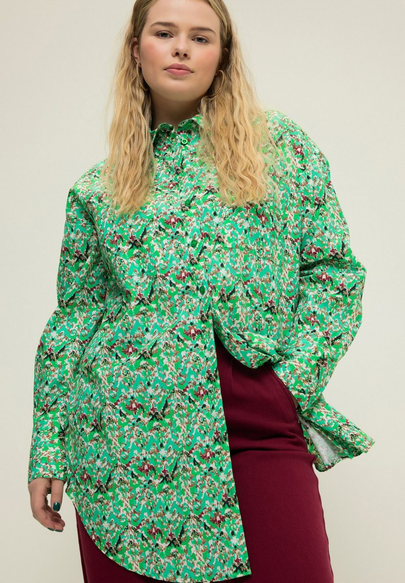 Blusa a maniche lunghe con colletto, verde a fantasia, e bottoni. Il design presenta forme astratte in tonalità di verde, rosso e nero.
