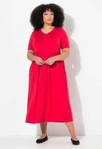 Robe rouge à manches courtes avec une taille froncée et un détail zippé. Le tissu est lisse avec une jupe fluide. Portée avec des chaussures plates noires.