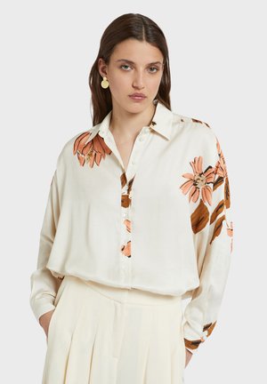 Kobieta w kremowej bluzce w pomarańczowe kwiatowe wzory i kremowej plisowanej spódnicy, ręce w kieszeniach, neutralny wyraz twarzy, rozpuszczone brązowe włosy.