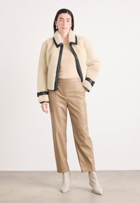 Faux shearling crèmekleurige jas met zwarte accenten, gecombineerd met tan leren broek. Model draagt een beige top en neutrale enkellaarsjes.