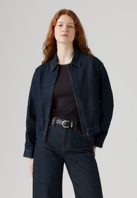 Veste en denim sombre avec un col, coupe décontractée et poignets élastiques. Portée sur un haut à côtes noir, associée à un pantalon en denim sombre et une ceinture.