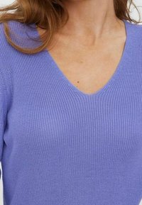 Vaaleanvioletti V-kaula-aukollinen neulepaita, jossa on teksturoitu kuvio. Kangas näyttää pehmeältä ja rentoilta, lyhyillä hihoilla.