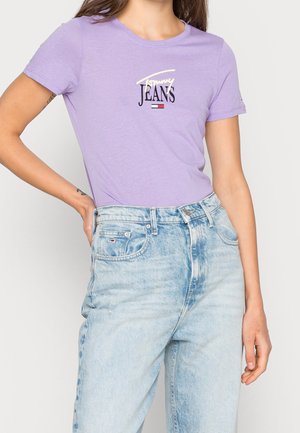 Woman wearing a light purple fitted T-shirt with "Tommy Jeans" logo and light blue high-waisted jeans, hand resting on hip.