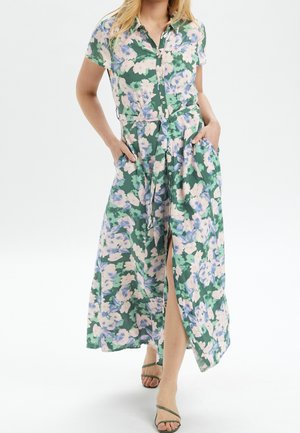 Kobieta ubrana w zielono-fioletową sukienkę na guziki z krótkimi rękawami i przednim rozcięciem, w połączeniu z zielonymi sandałami na paski, stoi.