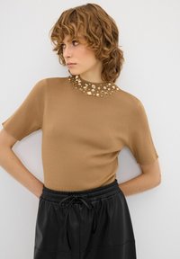Hnědý top s krátkým rukávem a vysokým krkem, zdobený průhlednými a zlatými ozdobami, kombinovaný s černými šortkami na stažení. Měkká textura.