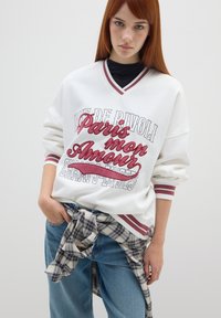 Felpa bianca oversize con testo grafico rosso e grigio. Presenta un collo a V e dettagli a righe sulle maniche. Jeanz in denim blu e camicia a quadri legata in vita.