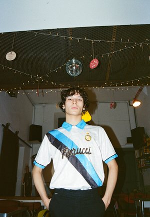 Witte blouse met blauwe en zwarte diagonale strepen, met het 'Fiorucci'-logo in bruin en een gouden ster boven een cirkelvormig embleem.