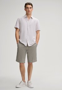 Camicia di lino leggera grigia a maniche corte con bottoni, abbinata a pantaloni corti grigi chiari con motivo e sneakers bianche. Il modello è in piedi con le mani nelle tasche.