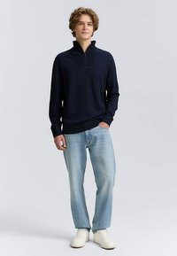 Tamsiai mėlynas megztinis su pusiau užtraukiamu apykakliu, derinamas su šviesiai mėlynais, tiesios kojų linijos džinsais ir baltais sportbačiais. Švelnios tekstūros, kasdieniškas dizainas.