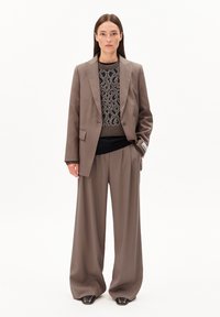 Vrouw in een taupe wijde broekpak en een sweater met luipaardpatroon, staande met één hand in de zak tegen een effen witte achtergrond.