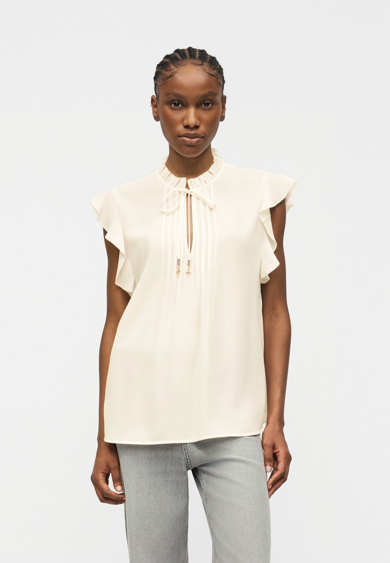 Femme aux cheveux tressés portant un chemisier sans manches crème avec des manches volantées et un col avec une ouverture nouée, associé à un jean gris clair.