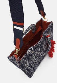 Textilní kabelka s vzorovaným vnějším povrchem v námořnické modři a krémové barvě, červenou látkovou podšívkou a červeným třásňovým akcentem. Obsahuje pevný námořnický popruh.