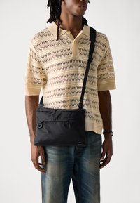 Svart nylon crossover-veske med frontlomme med glidelås, justerbar stropp, rektangulær form, glatt tekstur og minimalistisk design.
