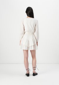 Una modella è di spalle, indossa un vestito chiaro a strati con vita arricciata e scarpe nere con lacci, su uno sfondo bianco semplice.