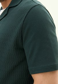 Tummansinisellä polo-paidalla on teksturoitu kuvio vartalolla, sileät hihat ja klassinen kaulus. Lähikuva sivusta.