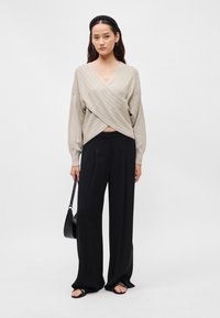 Pulover bej, texturat cu dungi diagonale, purtat cu pantaloni negri lejeri. Modelul ține o geantă neagră și poartă sandale plate.