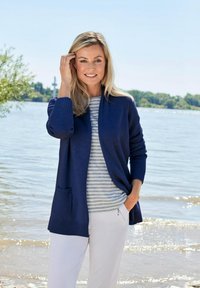 Cardigan bleu marine avec des poignets côtelés et des poches, porté sur une chemise rayée. Pantalons clairs, se tenant près d'un plan d'eau avec de la verdure en arrière-plan.