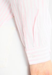 Camicia a maniche lunghe con righe verticali rosa e bianche, dettaglio dei polsini con bottoni, realizzata in un tessuto leggero. Presenta una vestibilità rilassata.