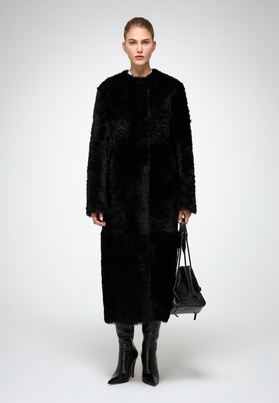 Cappotto in pelliccia sintetica nera con maniche lunghe, lunghezza ginocchio e superficie testurizzata. Indossato con stivali neri fino al ginocchio e tiene una borsa nera.