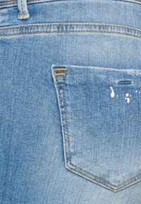 Jeans em denim azul claro com um bolso traseiro que apresenta costura amarela e detalhes em manchas de tinta branca. Tecido texturizado com uma aparência desgastada.