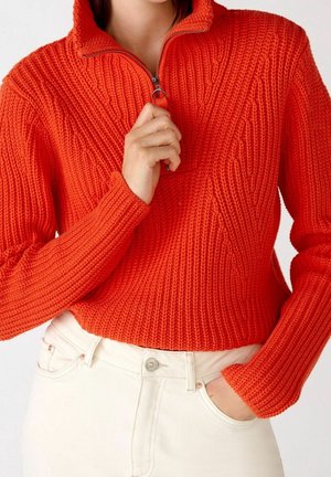 Kobieta ubrana w jaskrawo pomarańczowy, grubo dziergany sweter z półzamkiem przy kołnierzu, podciągająca ręką zamek do góry, w połączeniu z wysokim stanem białych spodni.