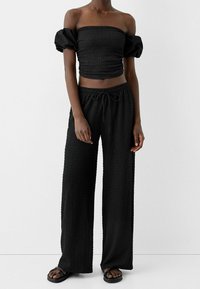 Ensemble noir deux pièces comprenant un haut sans bretelles texturé avec des manches bouffantes et un pantalon ample à cordon de serrage. Sandales complètent le look.