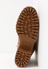 Semelle de botte marron avec des crampons profonds, présentant des motifs en forme de croix et rectangulaires pour la traction, posée sur un fond blanc.