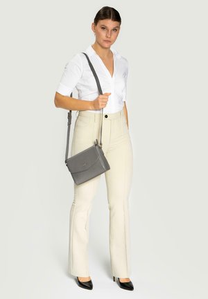 Graue, strukturierte Umhängetasche mit Klappenverschluss, getragen zu einem weißen Hemd mit Kragen und beigen, ausgestellten Hosen. Schwarze Absätze vervollständigen das Outfit.