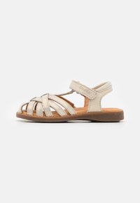 Sandalía de cuero beige con tiras entrecruzadas, cierre de Velcro y suela de goma marrón. Diseño de punta abierta con un acabado texturizado.