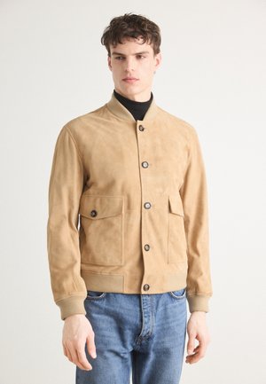 Khaki suede bomber jacket with ribbed collar and cuffs, featuring two front pockets and button closures, worn over a black turtleneck.