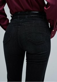 Los pantalones vaqueros de mezclilla negra presentan una cintura alta, dos bolsillos traseros con diseños angulares y una tela lisa y texturizada. La cinturilla tiene un acabado brillante.