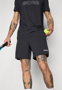Svart BORG treningsskjorte og shorts med teksturert stoff, holder en tennisball i den ene hånden og en racket i den andre.