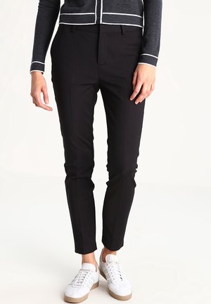 Person trägt enge schwarze Hose, weiße Sneakers und eine dunkelgraue Strickjacke mit weißen horizontalen Streifen und steht vor einem einfarbigen Hintergrund.