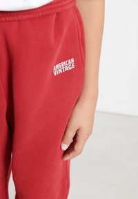 Raudoni medvilniniai džinsai su raukinėta tekstūra ir elastinga juosta. Šone yra baltas "AMERICAN VINTAGE" logotipas kišenės srityje.