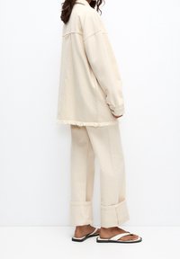 Giacca oversized beige con orlo sfrangiato, abbinata a pantaloni a gamba larga coordinati. Sandali semplici completano il look, mettendo in risalto una vestibilità rilassata.