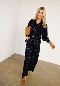 Χαμογελαστή γυναίκα με μακριά ξανθά μαλλιά φοράει μαύρο πουκάμισο με κουμπιά και παντελόνι φαρδύ με μικρές χρυσές βούλες, κρατώντας καφέ τσαντάκι, στέκεται σε τσιμεντένιο πάτωμα.