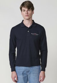 Tengerészkék hosszú ujjú póló gallérral, piros és fehér mellkasi logóval, textúrált anyagból. Világoskék farmernadrággal párosítva.